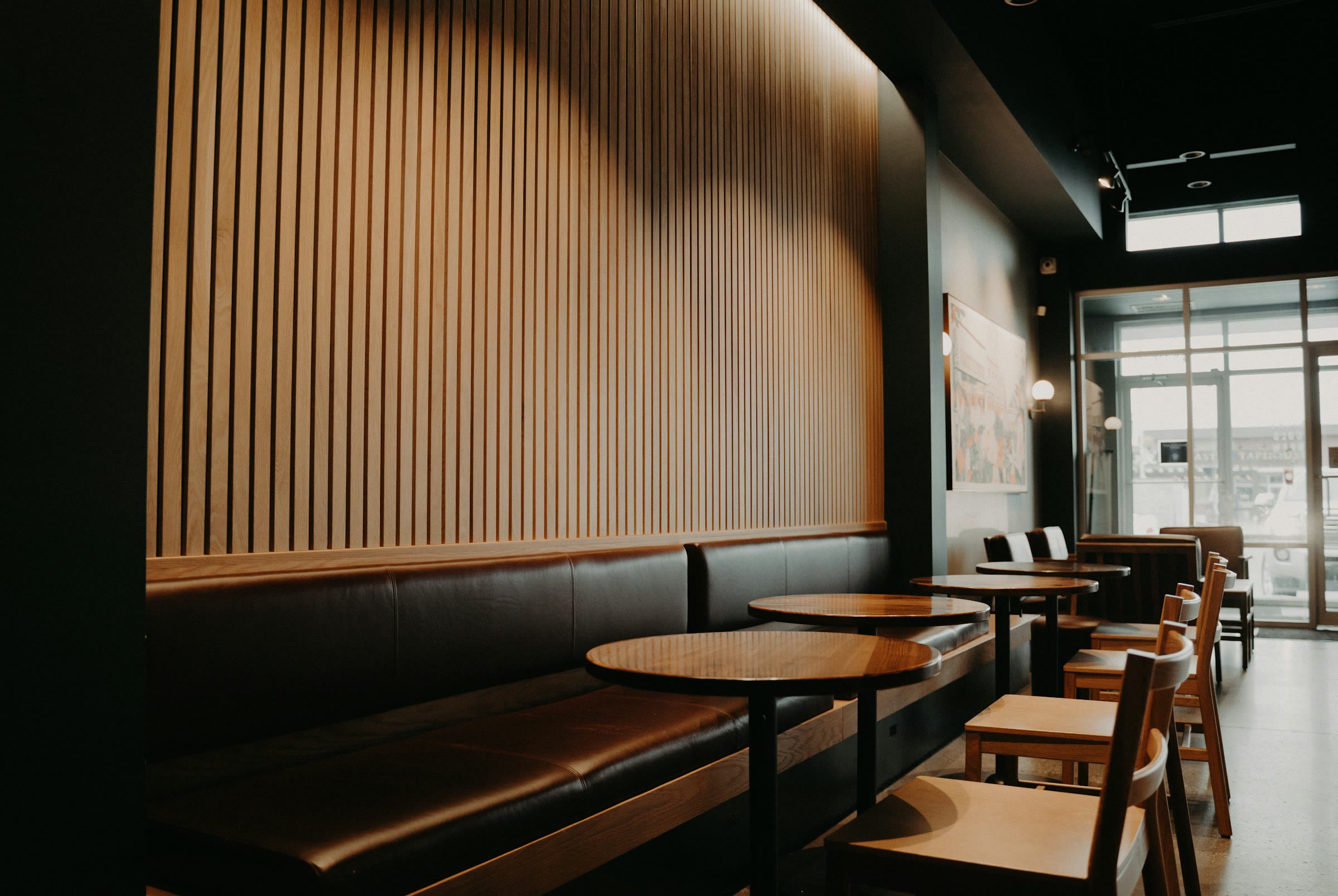 A dim room with a slatted timber wall, a long leather banquette and small round tables