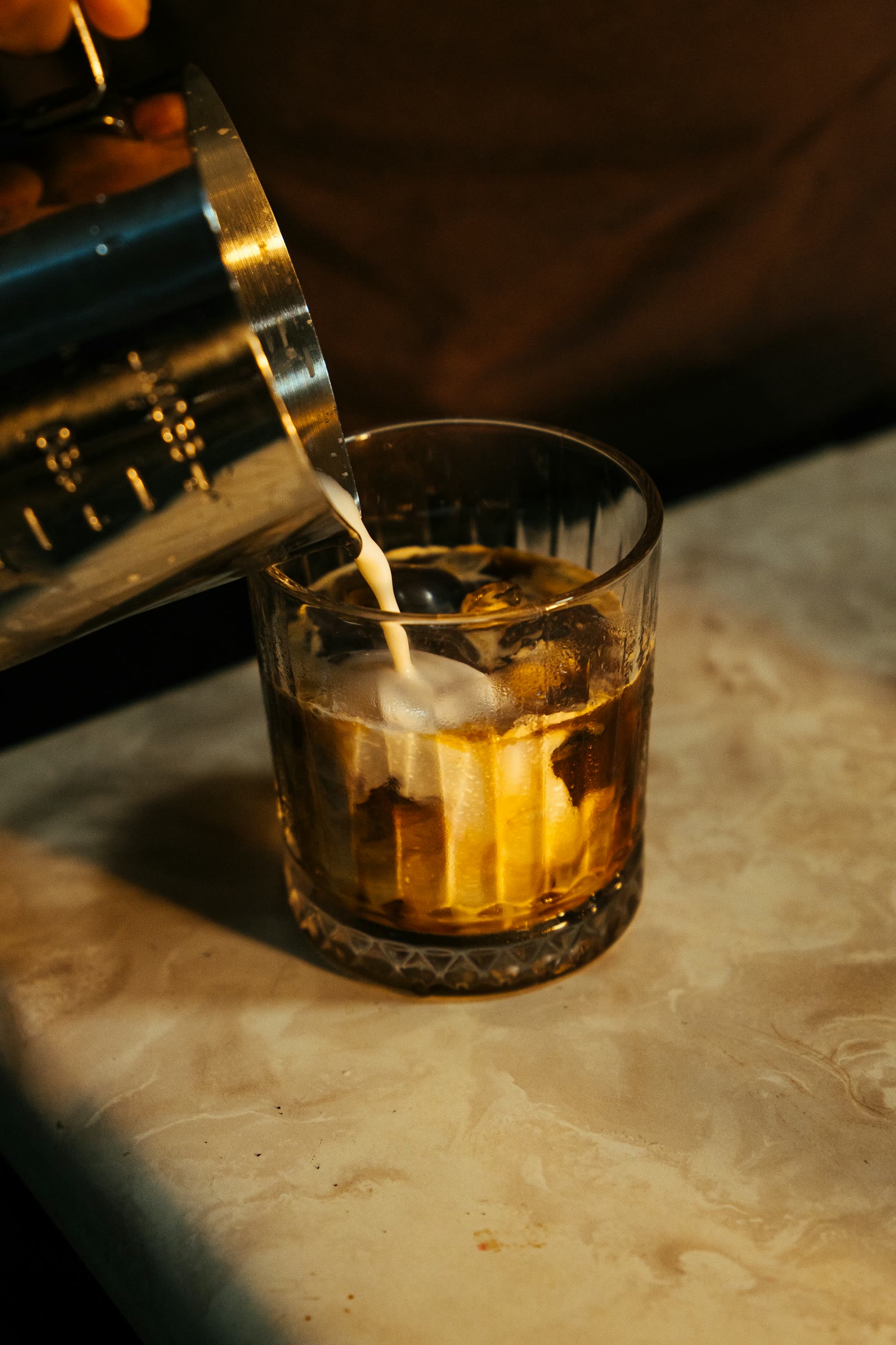 Milk poured from a steel pitcher into iced coffee on a marble counter