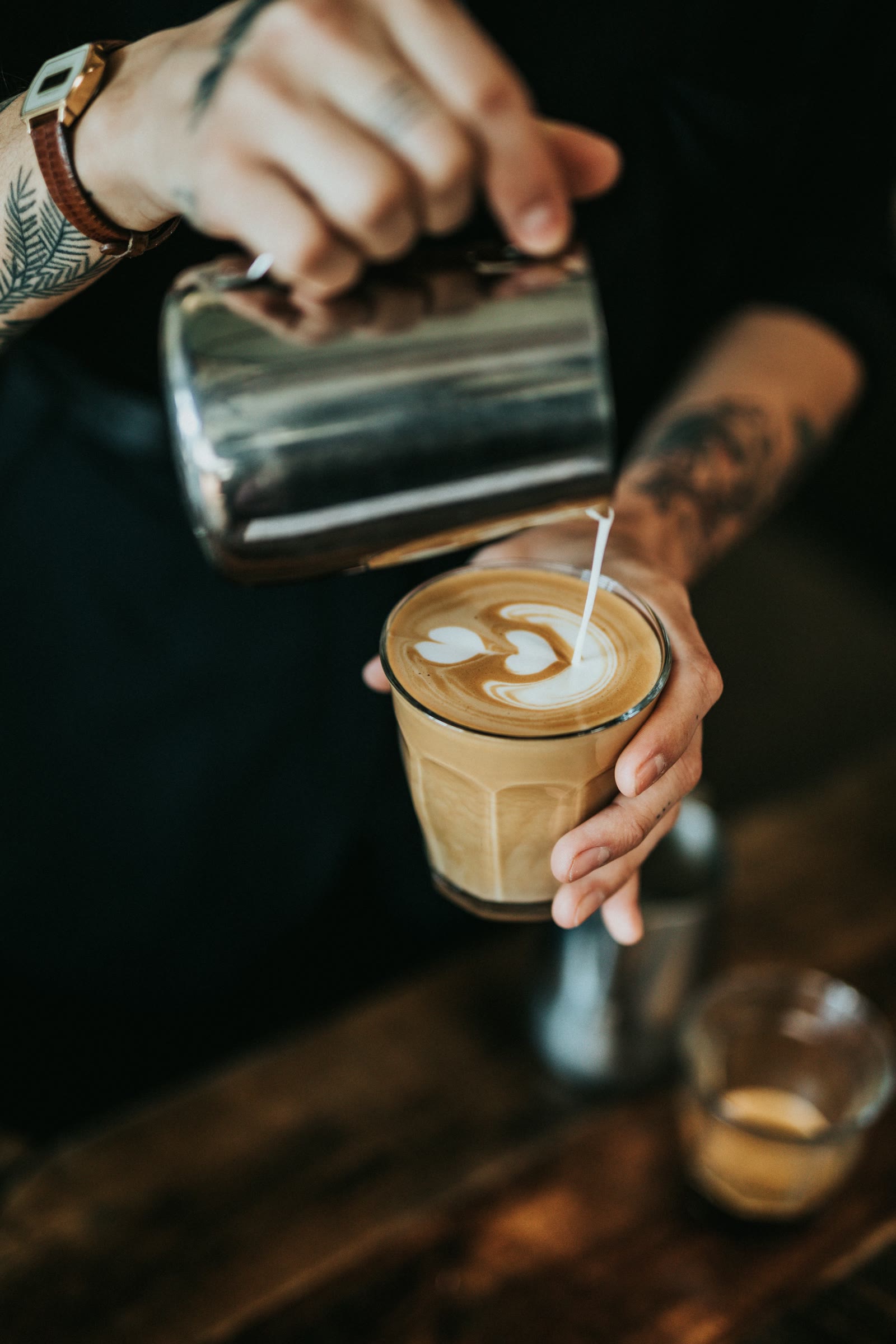A tattooed hand pouring steamed milk into a glass to draw a rosetta