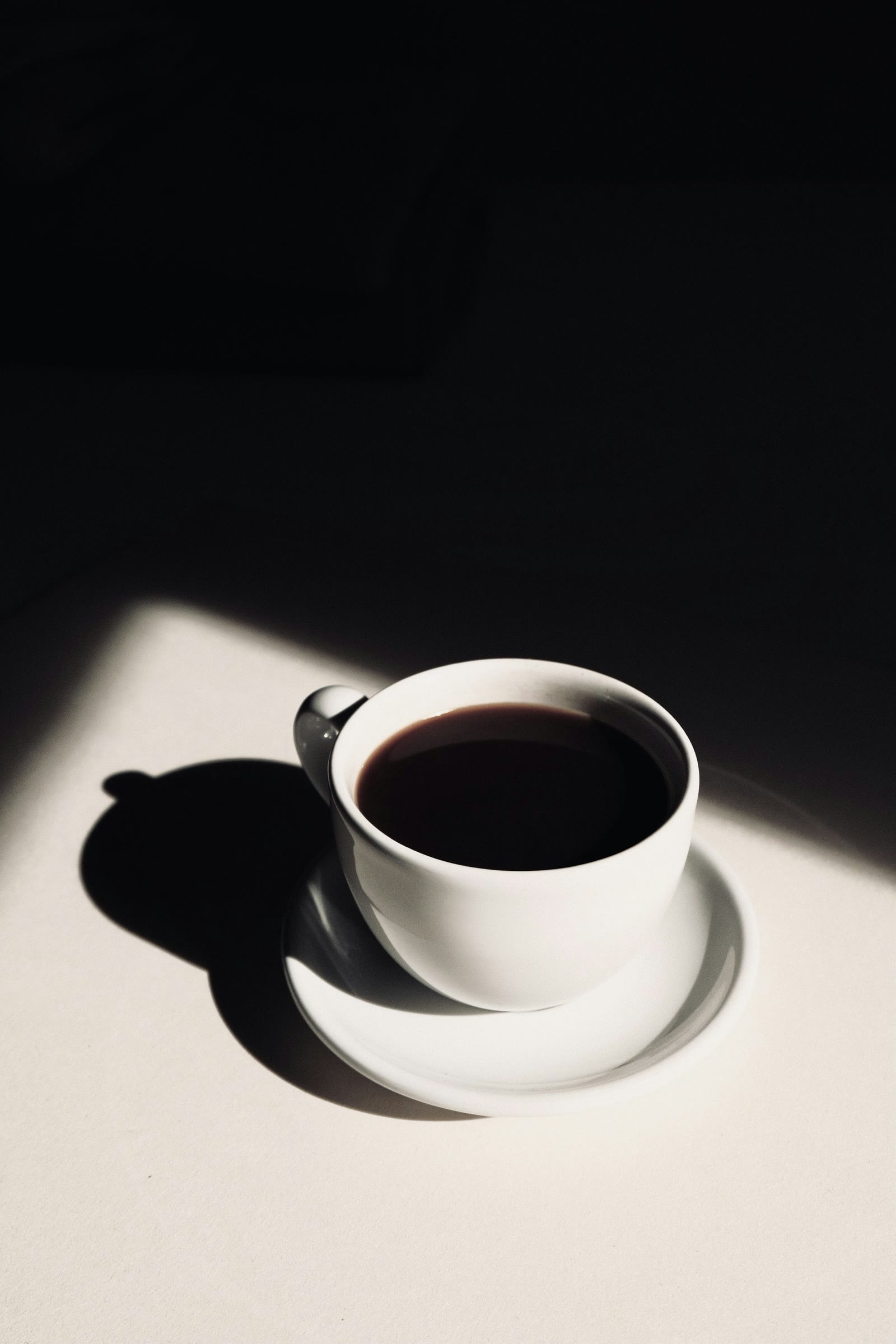 A white cup of black coffee on its saucer, half caught in a hard-edged patch of sunlight